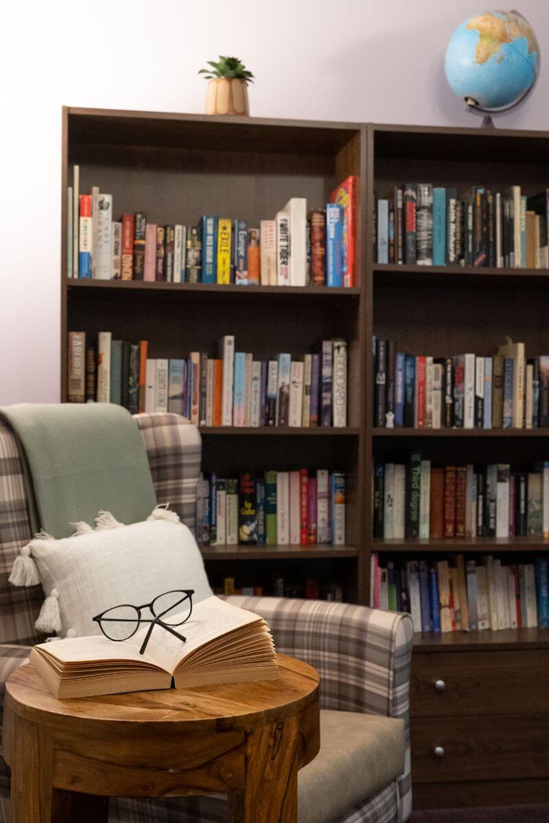 A bookcase with globe on top, glasses on a open book and a checked armchair.