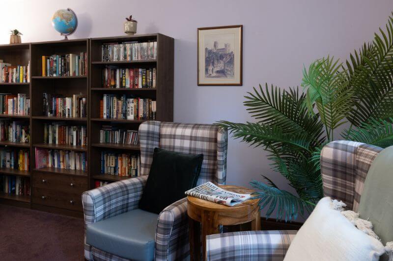Two checked armchairs with a plotted plant and a coffee table with newspaper on top and next to a bookcase.