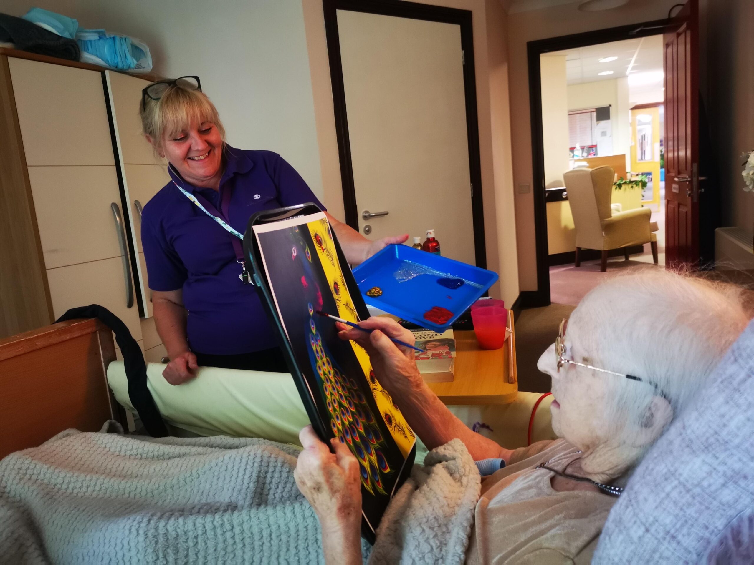 A female resident in bed painting with help of a female carer who is smiling.