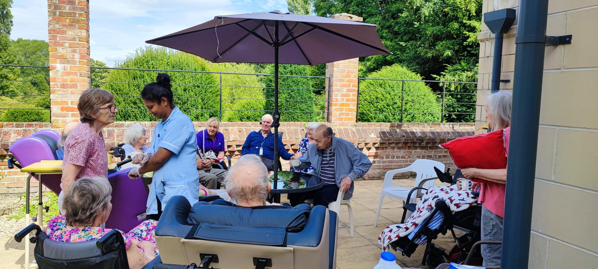 Residents in the garden having a cup of tea under a umbrella. A carer helping a resident.