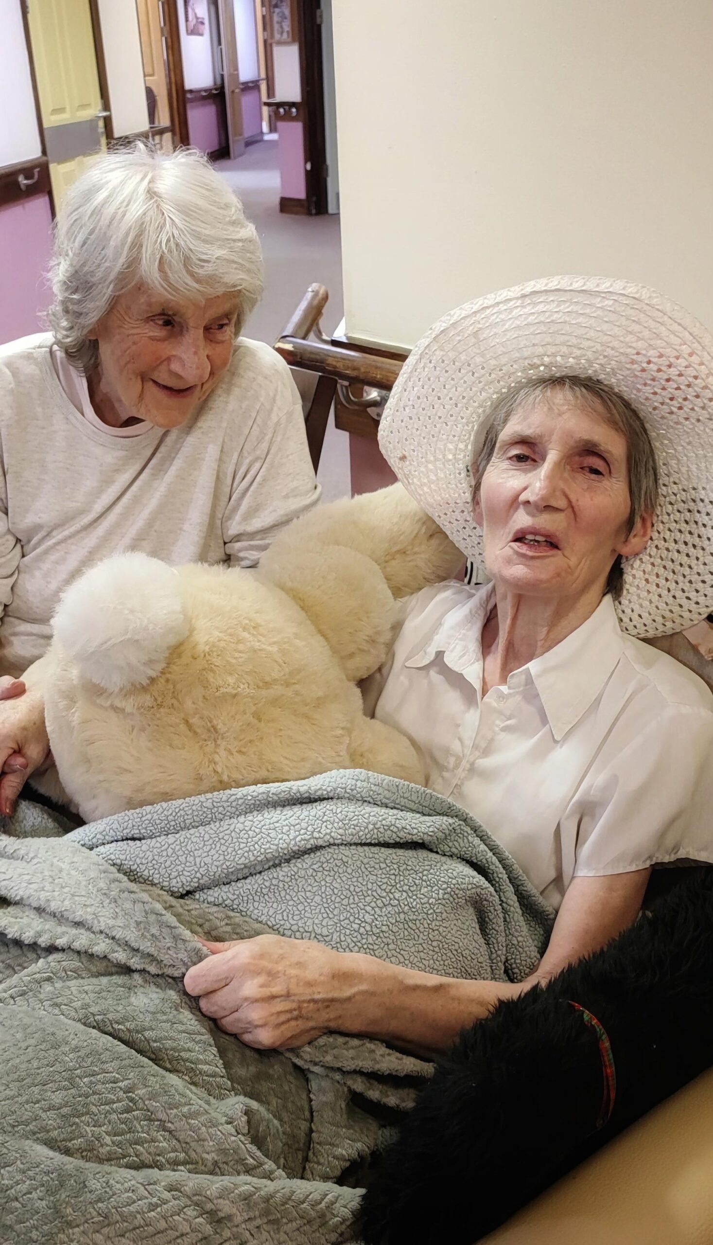 Two elderly residents smiling. One looking at the other and the other looking at the camera under a blanket wearing a hat.