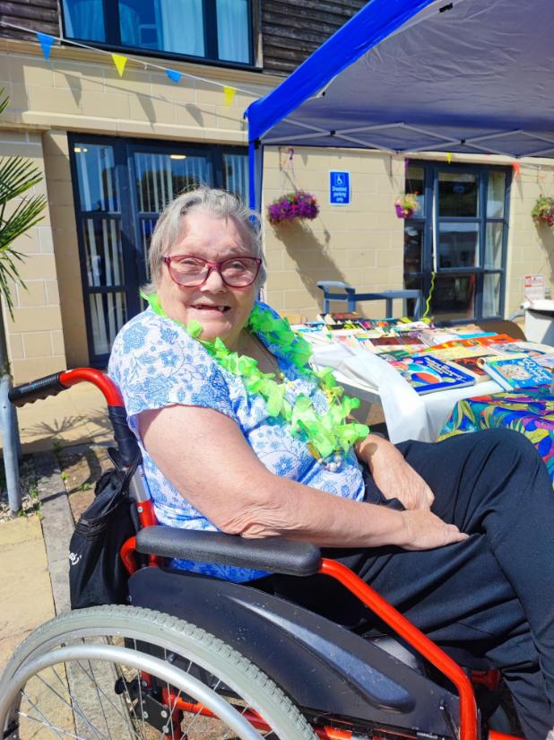 older lady with a green flower necklace sat in a wheelchair smiling